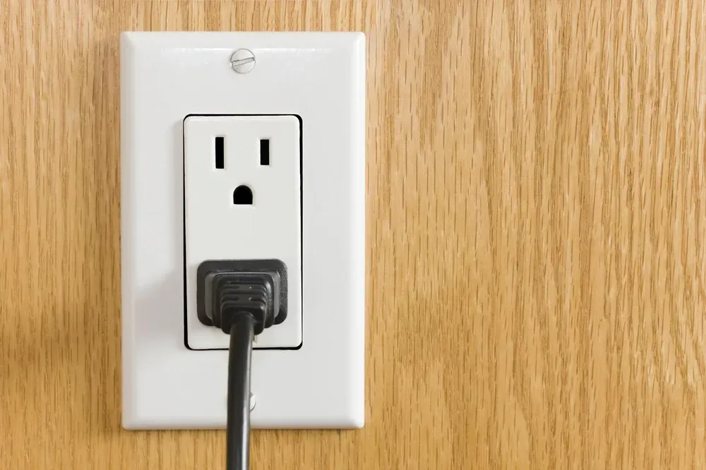 White duplex wall outlet with a black plug on a wood-grain wall — does NOT show a ceiling fan