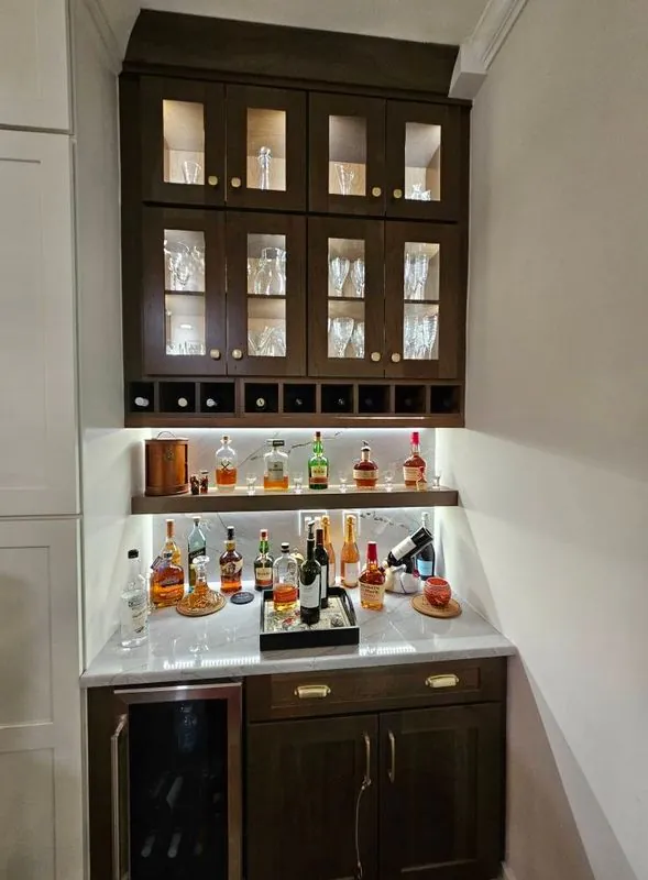 Home bar nook: dark wood glass-front upper cabinets with in-cabinet lighting, lit shelf of bottles, wine fridg